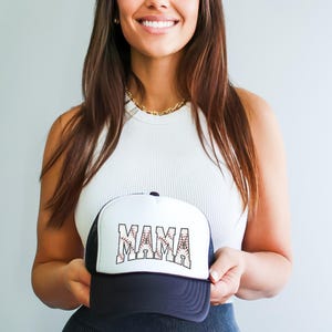 May include: A white and black baseball cap with a mesh back. The front of the cap has the word "MAMA" written in black letters with a baseball stitch design.