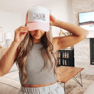 May include: A pink and white trucker hat with the word "MAMA" in black lettering. The hat is worn by a person with long blonde hair.