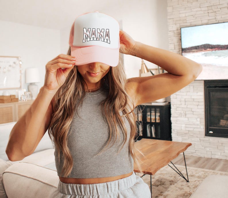May include: A pink and white trucker hat with the word "MAMA" in black lettering. The hat is worn by a person with long blonde hair.