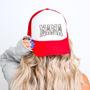 May include: A white and red trucker hat with a baseball-themed "MAMA" graphic. The hat has a mesh back and a curved brim.