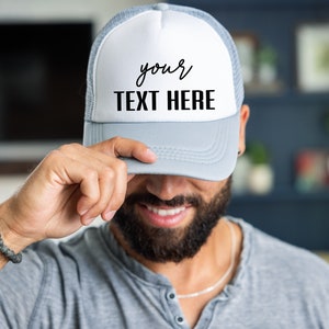 May include: A gray and white trucker hat with a white mesh back. The front of the hat has black text that says "your TEXT HERE".