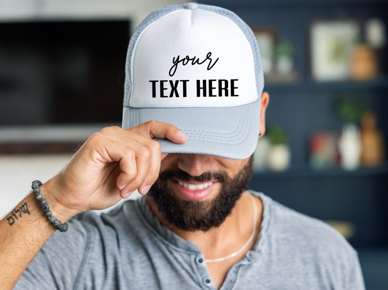 May include: A gray and white trucker hat with a white mesh back. The front of the hat has black text that says "your TEXT HERE".