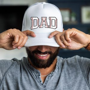 May include: White trucker hat with the word "DAD" in a baseball-themed design. The letters are outlined in black with a baseball stitch pattern. The hat has a mesh back and a curved brim.