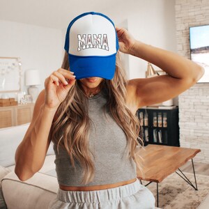 May include: A blue and white trucker hat with the word "MAMA" in white letters with a red baseball stitch design. The hat is being worn by a person with long blonde hair.