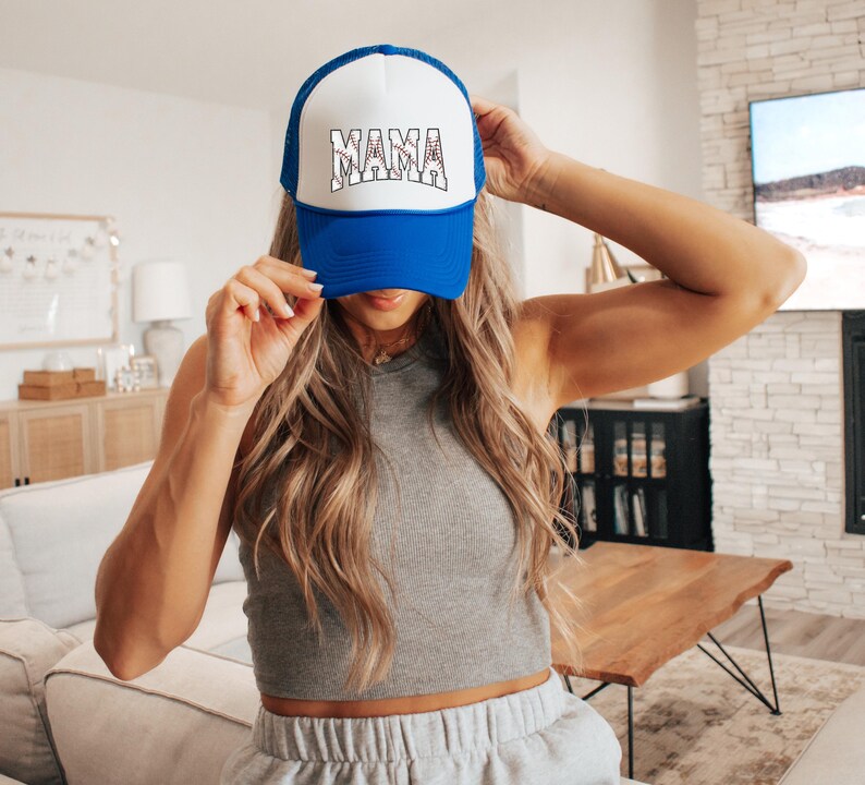 May include: A blue and white trucker hat with the word "MAMA" in white letters with a red baseball stitch design. The hat is being worn by a person with long blonde hair.