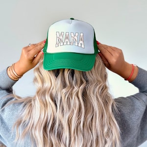 May include: Green and white baseball cap with a white mesh back. The front of the cap has the word "MAMA" written in white letters with a baseball stitch design.