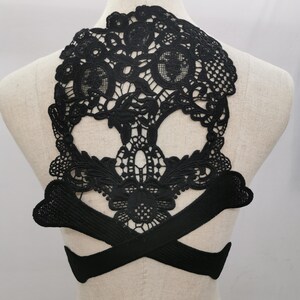 May include: Black lace skull applique with crossbones. The intricate design features a detailed skull shape with open eye sockets and a delicate floral pattern. The crossbones are positioned below the skull.