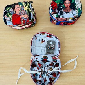 May include: Three round fabric jewellery cases featuring colourful prints of Frida Kahlo. Two cases are closed with zips, while one is open, revealing a white interior lining with a red heart pattern, a drawstring pouch, and small earring storage.