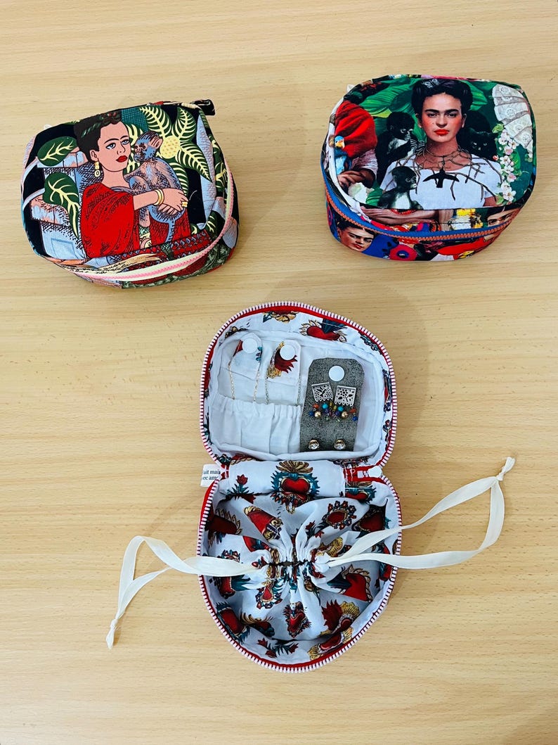May include: Three round fabric jewellery cases featuring colourful prints of Frida Kahlo. Two cases are closed with zips, while one is open, revealing a white interior lining with a red heart pattern, a drawstring pouch, and small earring storage.