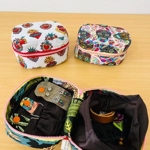 May include: Two small, rectangular zipped pouches with colourful prints: one featuring red sacred hearts on white, the other with multi-coloured sugar skulls. An open pouch displays a black interior with a small organiser flap and a drawstring compartment.