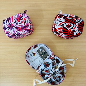 May include: Two small, square, zipped fabric pouches featuring colourful floral and animal patterns in purple, pink, red, and black. A third, open, round zipped pouch displays an interior with white fabric, small pockets, and a drawstring compartment.