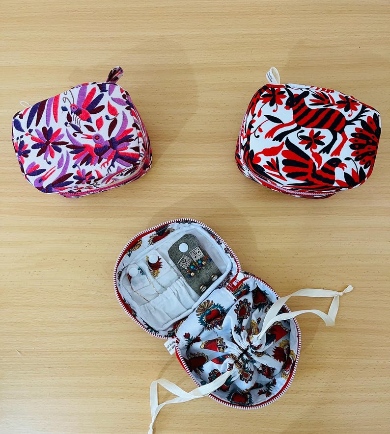 May include: Two small, square, zipped fabric pouches featuring colourful floral and animal patterns in purple, pink, red, and black. A third, open, round zipped pouch displays an interior with white fabric, small pockets, and a drawstring compartment.