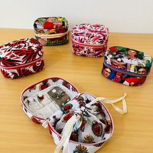 May include: Five small, rectangular, zipped fabric cases with various colourful prints, including floral patterns and portraits. One case is open, revealing a white interior with a drawstring pouch and small compartments for jewellery storage.