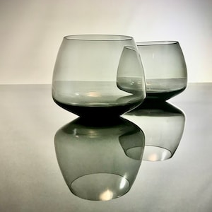 May include: Two clear glass tumblers with a smoky gray tint. The tumblers are round and have a wide base. The larger tumbler is in the foreground and the smaller tumbler is behind it.
