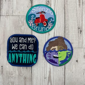 May include: Three embroidered patches. One is round and blue with a red scooter and the text "Silenzio Bruno". Another is rectangular and blue with the text "You and me? We can do anything". The last is round and blue with a cartoon face with half of it underwater.