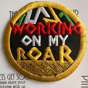 May include: A circular embroidered patch with a black background and a thick yellow border. The text "WORKING ON MY ROAR" is stitched in red, white, and yellow thread. Above the text are green and yellow shapes, and below is a geometric pattern.