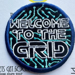 May include: A round, embroidered patch with the text "WELCOME TO THE GRID" in white, outlined in black, on a black background. The patch has a blue border and teal accents. The patch is likely for clothing or accessories.