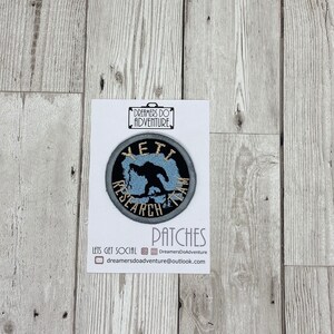 May include: A round, grey and blue embroidered patch with the text "YETI RESEARCH TEAM" in gold lettering. The patch features a silhouette of a Yeti in the centre.