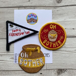 May include: Three embroidered iron-on patches. One is a red and yellow circle with the text "Oh bother" and a honey pot. Another is a brown honey pot with the text "Oh bother". The last is a white and black triangle with the text "Happy Friendship Day".