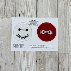 May include: Two embroidered patches on a white card. One patch is white with a black line and dots, and the text "BAH-LA-LA-LA". The other is red with a white face design. The card reads "PATCHES" and "Dreamers Do Adventure".