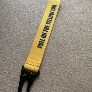 May include: A bright yellow fabric strap featuring the black text PULL ON THE YELLOW TAG printed vertically. The strap is securely attached to a black metal carabiner clip, providing a functional attachment point for keys or other small items.