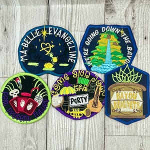 May include: Five embroidered patches with various designs and text. The patches are on a wooden surface. The patches include "Ma Belle Evangeline", "We're Going Down The Bayou", "Come And Join The Party", "Past Present Future", and "Bayou Beignets".