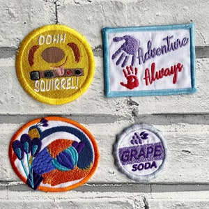 May include: Four embroidered patches with different designs. One patch is yellow with a cartoon squirrel and the text "Oohh Squirrel!" Another patch is blue with a purple handprint and the text "Adventure Always". A third patch is orange with a blue and purple flower design. The final patch is white with a purple grape design and the text "Grape Soda".