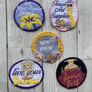 May include: Five embroidered iron-on patches featuring characters and quotes from the Disney movie Tangled. The patches are circular and oval in shape and feature a purple, yellow, pink, white, and black background. The text on the patches reads: "Let your power shine", "Tower Art Supplies", "Who knew", "Go live your dreams", and "Mother knows best".