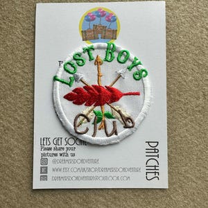May include: Embroidered patch with the text "Lost Boys Club" in green, featuring arrows and a red feather design. The patch is on a white background with a stitched border, and the card also includes social media information.