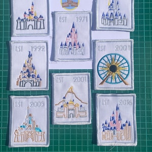May include: Nine white embroidered patches with different designs and dates. The patches feature a castle, a Ferris wheel, and a mountain. The dates on the patches range from 1955 to 2016.