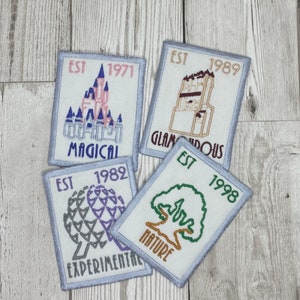 May include: Four embroidered patches with light blue borders. Each patch features a different design and text: a castle with "MAGICAL" and "EST 1971", a building with "GLAMOROUS" and "EST 1989", a geometric shape with "EXPERIMENTAL" and "EST 1982", and a tree with "NATURE" and "EST 1998".