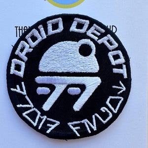 May include: Black circular patch with white embroidered text and design. The text reads "DROID DEPOT" around the top and "2017 FMUD" around the bottom. The central design is a stylized droid head and body.