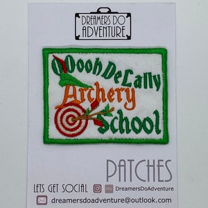 May include: A green, white, and orange embroidered patch with the text "Oooh De Lally Archery School". The patch features a target with an arrow through it.