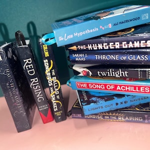 May include: A collection of miniature book replicas featuring titles such as The Love Hypothesis, The Hunger Games, Throne of Glass, Twilight, The Song of Achilles, Lights Out, Sunrise on the Reaping, Red Rising, and Haunting Adeline with ribbon pulls.