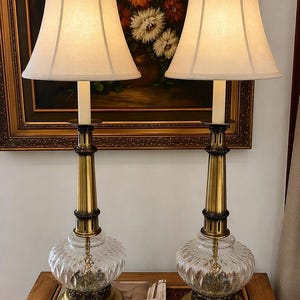 May include: Two vintage table lamps with cream-colored shades. The lamps have a brass finish with a clear, patterned glass base. The lamps are placed on a wooden table in front of a framed painting with floral imagery.