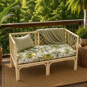 Könnte beinhalten: Ein helles Rattan-Sofa mit einem rautenförmigen Rahmen, einem Sitzkissen mit grün-blauem botanischem Blumenmuster, einem einfarbigen hellgrünen rechteckigen Zierkissen und einer darübergelegten salbeigrünen Strickdecke.