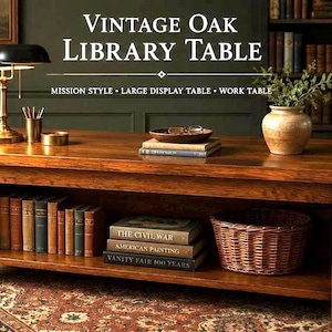 May include: A rectangular vintage oak library table with a warm brown wood grain finish. The table features a lower shelf holding a row of hardcover books and a woven wicker basket. The top surface displays a lamp, books, and a ceramic vase.