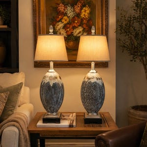 Puede incluir: Dos lámparas de mesa decorativas con bases ovaladas en azul y blanco. Cada lámpara tiene una pantalla beige y un remate de cristal. Las lámparas están sobre una mesa de madera frente a una pintura.