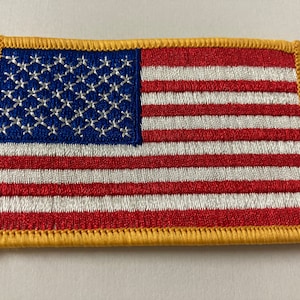 Puede incluir: Un pequeño parche bordado de la bandera estadounidense con un borde dorado. La bandera es roja, blanca y azul con 13 rayas y 50 estrellas.