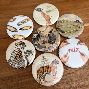 May include: Seven circular pinback buttons featuring diverse designs including a football stadium, a tiger, makeup supplies, ballet flats with Mizzou text, a tiger with a champagne tower, and hands holding lipsticks with the text miz and go tigers!