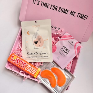 May include: A pink gift box containing self-care products. Inside are a facial mask, eye patches, a chocolate bar, and a pink chai tea packet. The facial mask is labelled "Radiate Love". The box is filled with pink shredded paper.