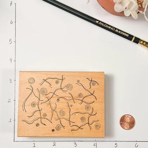 May include: A rectangular wooden stamp with an abstract design of swirling lines and circles. The stamp is approximately 3 inches long. A pencil and a penny are next to the stamp.