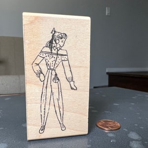 May include: A wooden stamp with a black and white illustration of a person wearing a patterned outfit and a headband. The person is standing with their arms at their sides and their feet slightly apart.