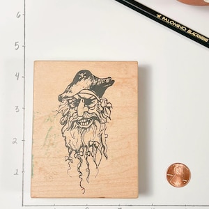 May include: A wooden stamp featuring a detailed illustration of a pirate's face with an eye patch, a hat, and a long beard. The stamp is rectangular and made of light-colored wood. A penny is next to the stamp for size comparison.
