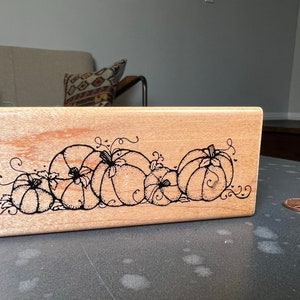 May include: A wooden stamp with a black and white design of pumpkins and vines. The stamp is rectangular and has a light brown color.