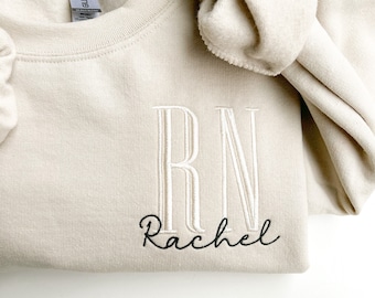 Könnte beinhalten: Ein beiger Pullover mit den Buchstaben "RN" in weiß gestickt und dem Namen "Rachel" in schwarz darunter.