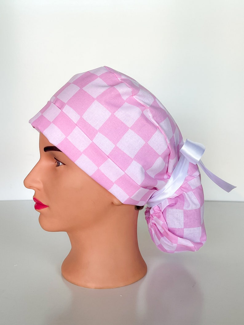 May include: Pink and white checkered surgical scrub hat with a white tie. The hat has a ponytail hole in the back.