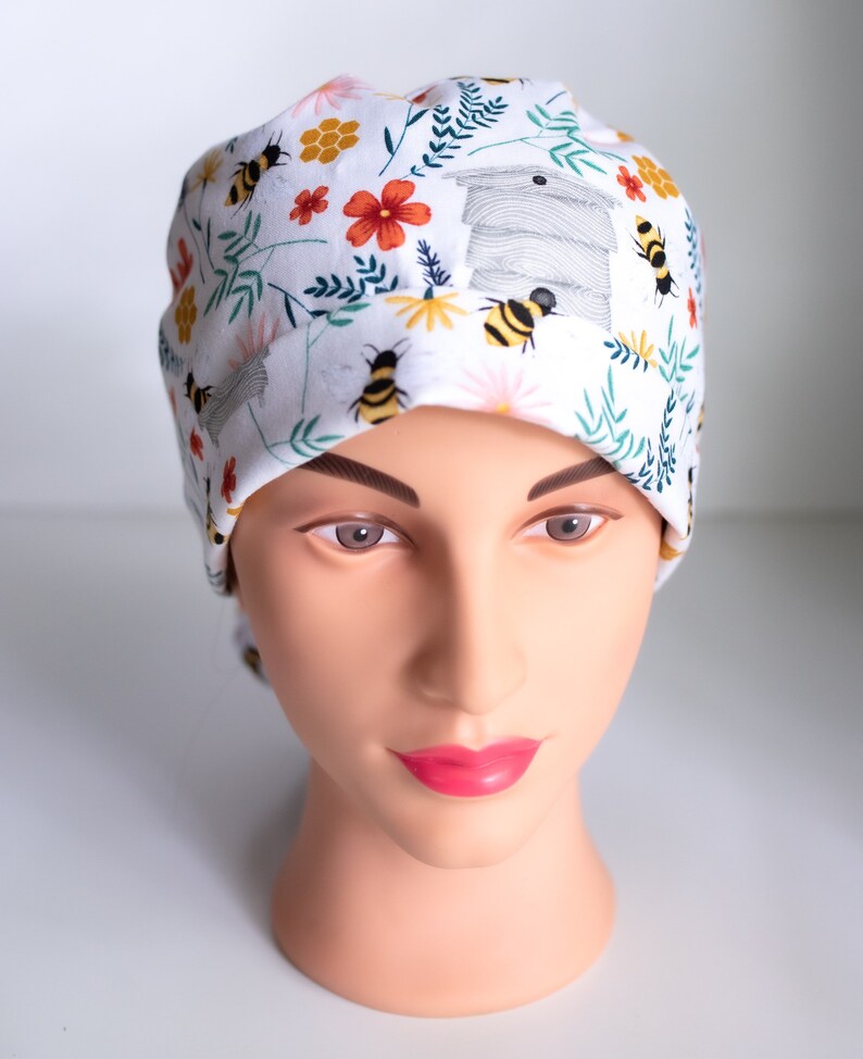 May include: A white scrub hat with a floral pattern featuring bees, flowers, and a honeycomb design.