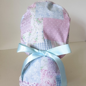 May include: A scrub cap featuring a patchwork design of light blue and pink floral, gingham, and heart patterns. The cap is secured at the back with a smooth, light blue satin ribbon tied into a bow, creating a gathered and adjustable fit.