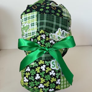 May include: A patchwork scrub cap with a green satin ribbon bow. The cap features various green patterns, including plaid, shamrocks, and the words "Happy St." in white. The cap is designed for medical professionals or those in the healthcare field.